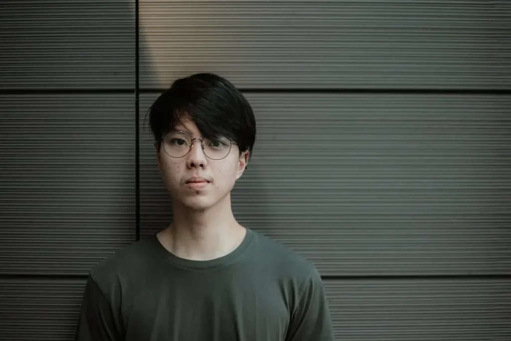 Portrait of a young asian professional with glasses wearing a fitted dark green t-shirt against a grey wall.
