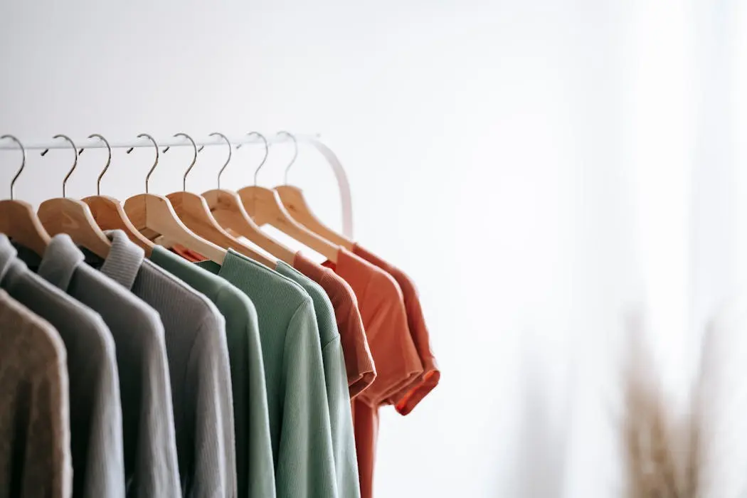 Clothing rack filled with t-shirts in various earth tones (sage green, grey, terracotta) on white hangers, representing fabric options.