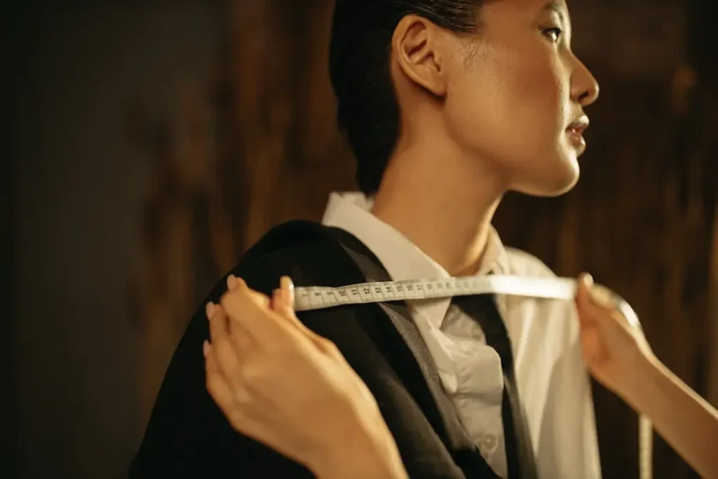 Custom Uniform Measurement Service | JS Uniform KL - T-Shirt Printing KL | Custom Shirt Printing Malaysia Professional tailor taking shoulder measurements of a model wearing a black suit jacket using a white tape measure.