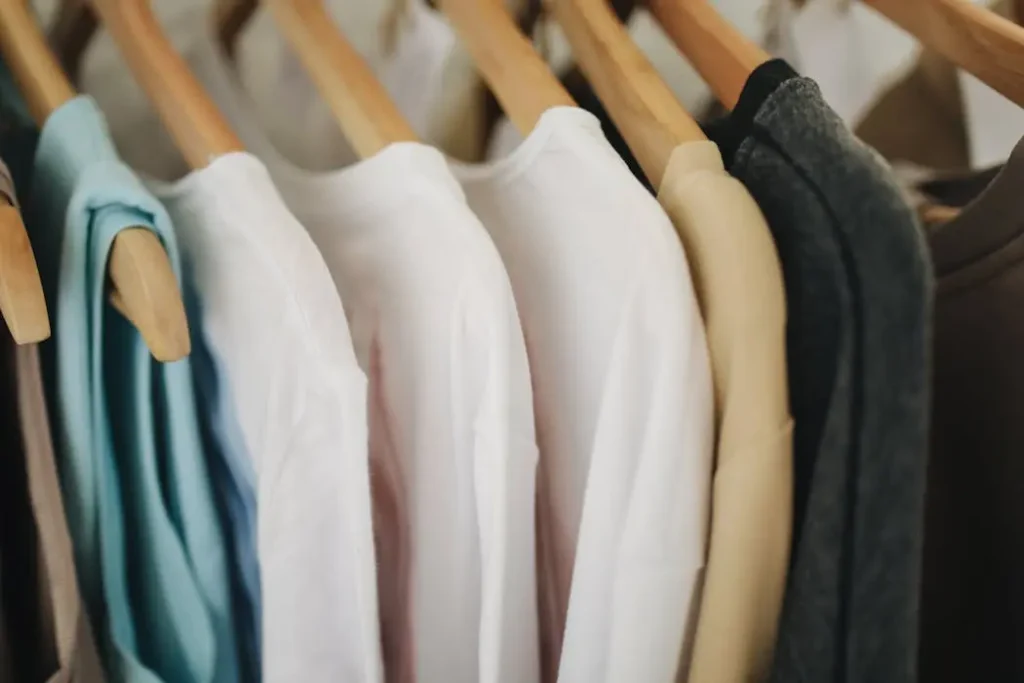 Close-up detail of soft, neutral-colored t-shirts hanging on a wooden rack, highlighting the fabric texture and quality.