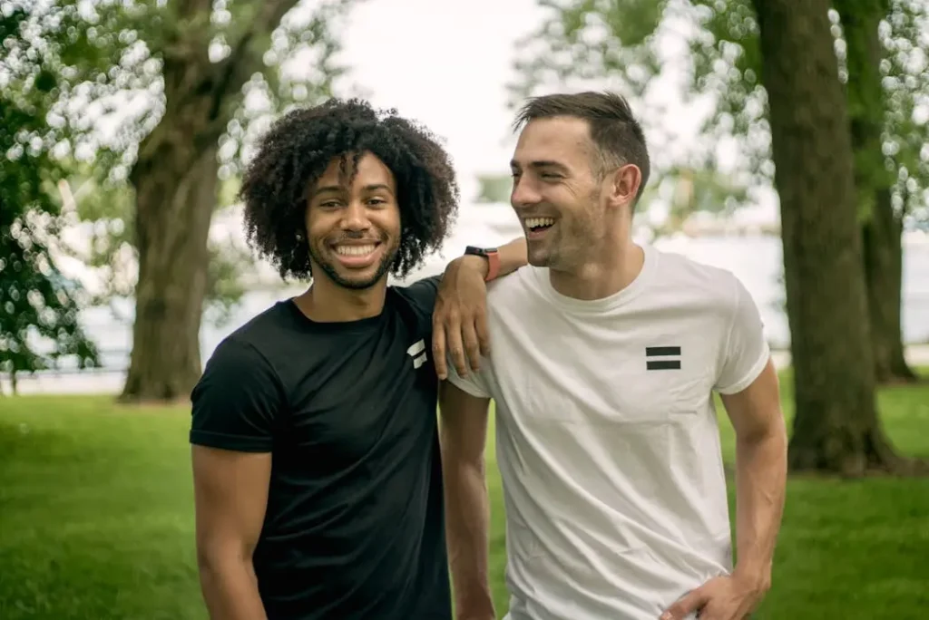 Two men smiling and embracing, wearing matching black and white t-shirts with a simple minimalist bar logo.