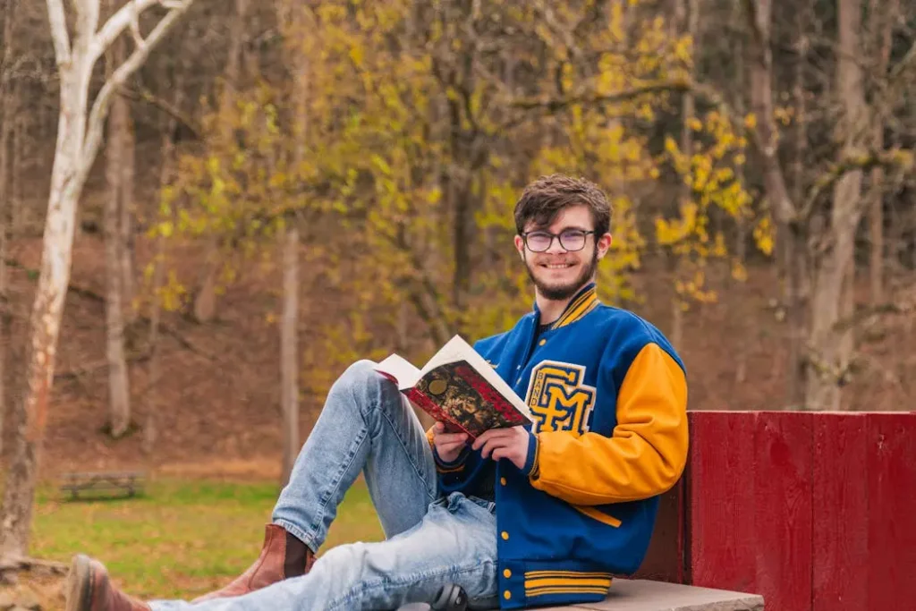 Student sitting outdoors reading a book, wearing a blue and yellow custom varsity jacket with a chenille patch letter logo.