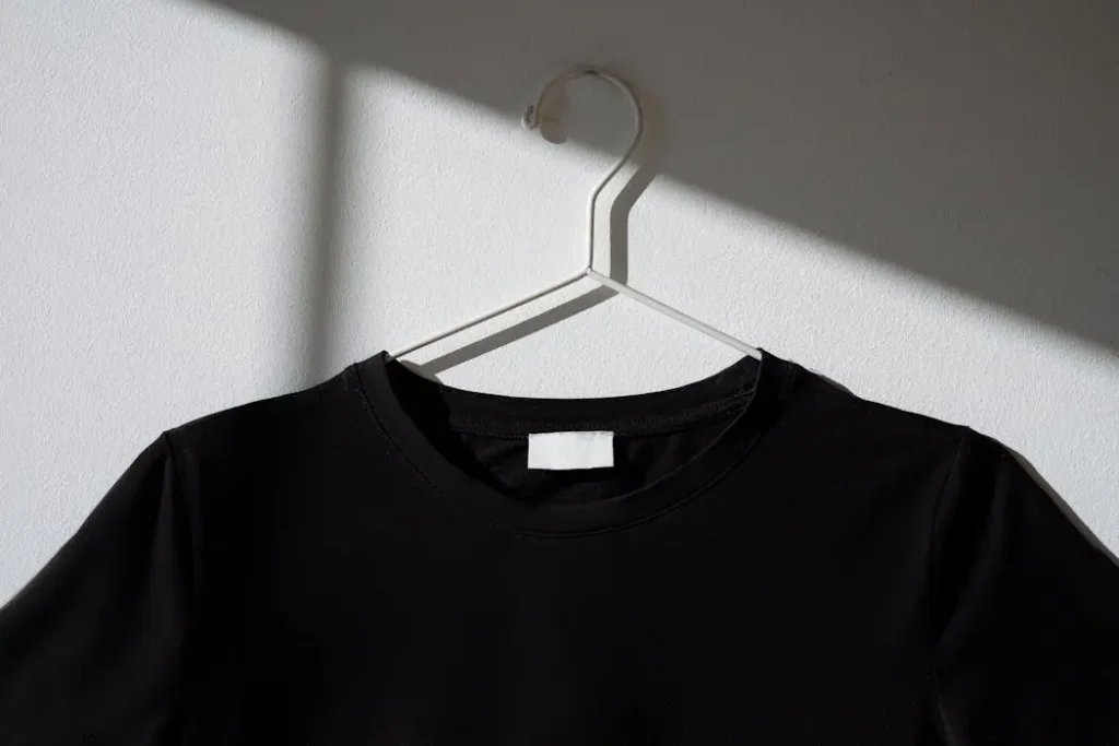 Minimalist black t-shirt hanging on a white wire hanger against a white wall, representing the premium, textured finish of embroidered apparel.