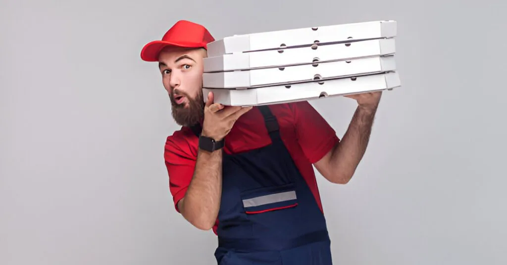 Pizza delivery man with a beard wearing a red cap and matching uniform holding a stack of white pizza boxes.