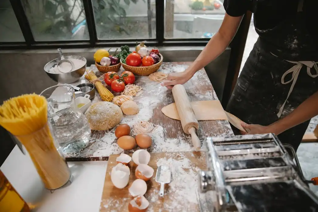 A chef wearing a premium custom-made black apron using a rolling pin to flatten dough on a floured wooden table, surrounded by fresh tomatoes, eggs, and a pasta maker.