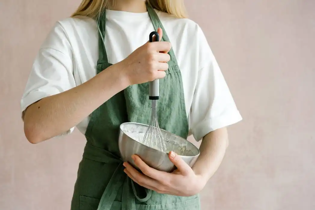 A cafe staff member wearing a white t-shirt and a light green custom-made apron, holding a stainless steel bowl and whisking ingredients.